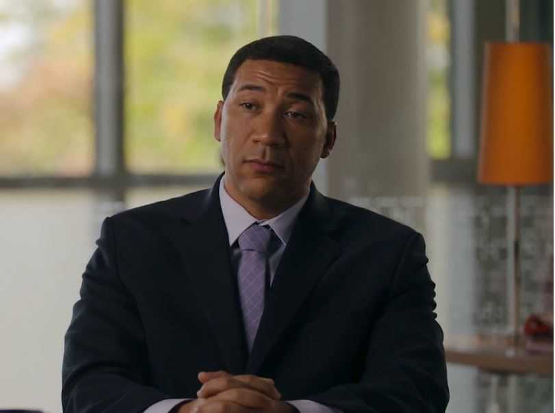 Man sitting with dark jacket and purple tie.